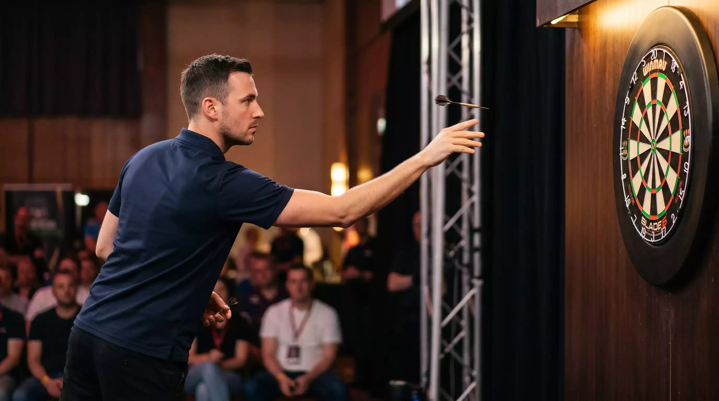 Darts-Spieler am Oche während eines WM-Matches mit Anzeigetafel im Hintergrund